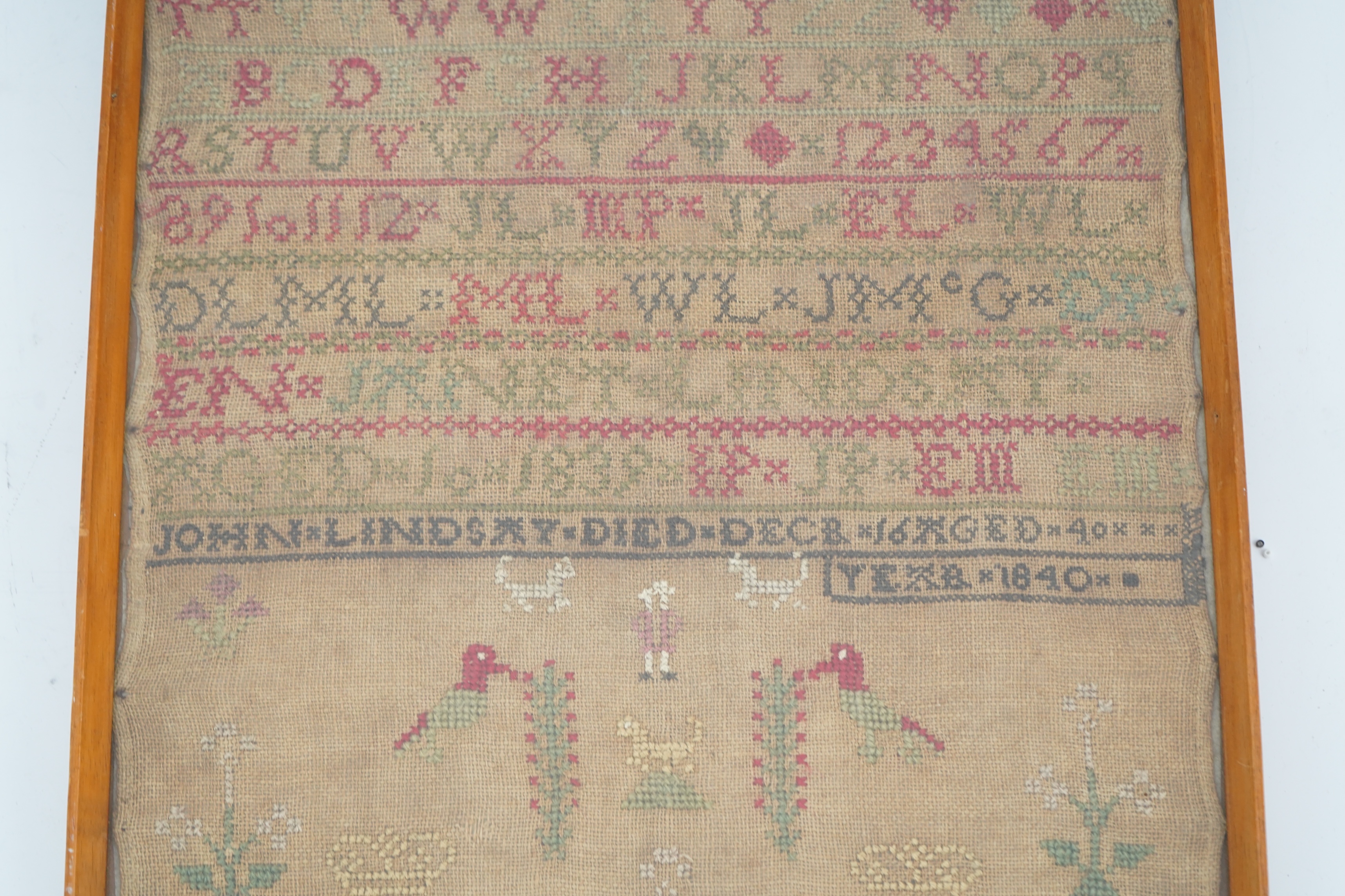 An alphabet cross stitch sampler with a figurative lower spot motif panel and commemorative dedication to John Lindsay, died Dec 16 1840, 27cm wide x 43.5 cm high. Condition - some light staining to linen, the blue and g